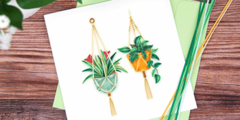 A white square greeting card with quilling botanical themed designs on the front in hanging pots with a green envelope laying on a dark wooden surface